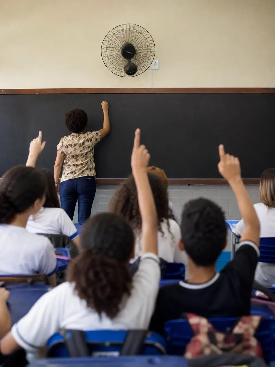 Professor escrevendo no quadro enquanto alunos levantam a mão