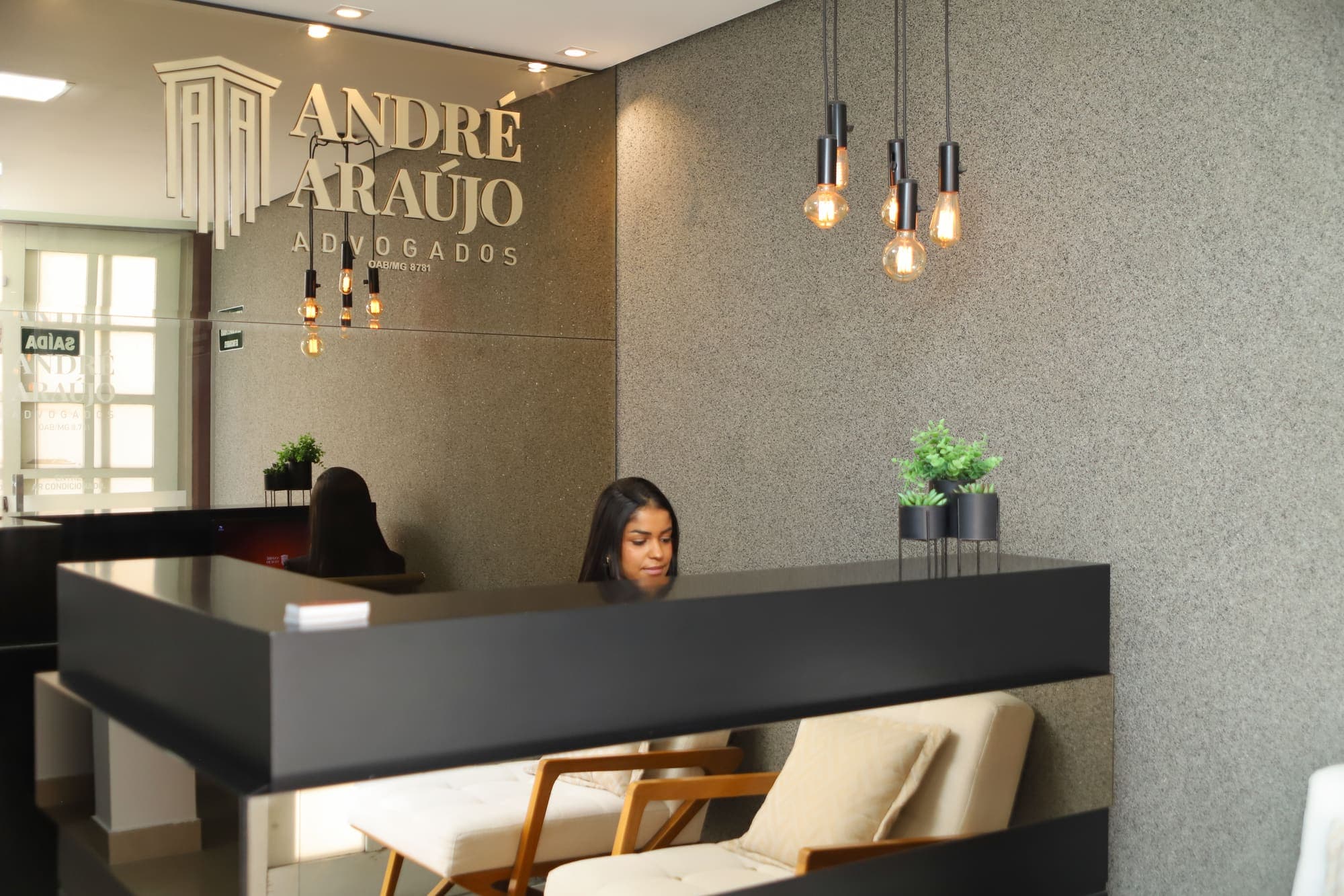 Recepcionista no balcão da recepção, sob o logotipo dourado e as luminárias de filamento
