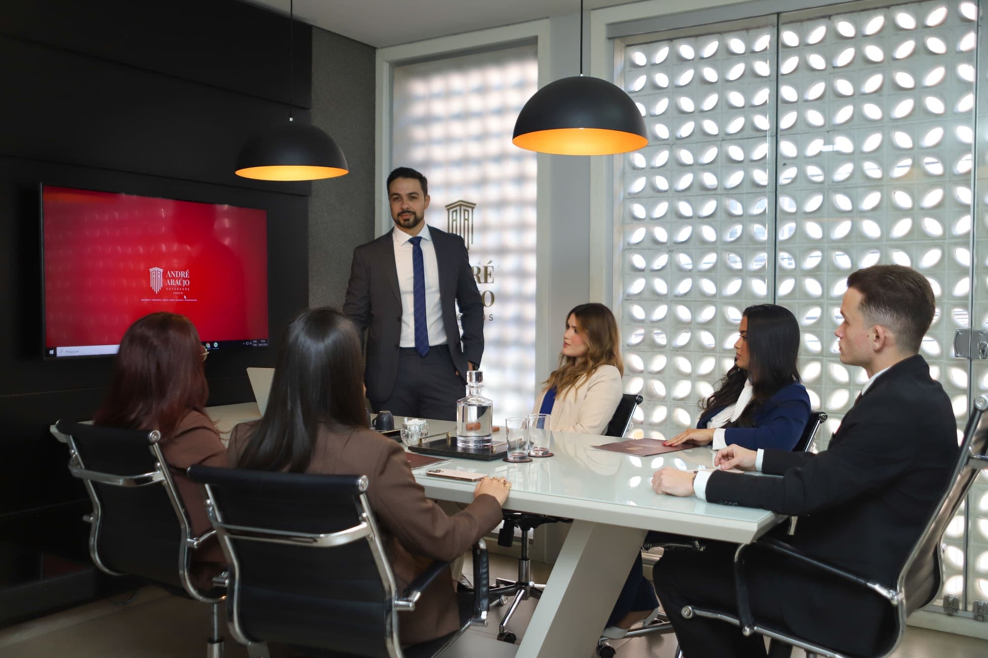Reunião em andamento na sala de reuniões, com a equipe à mesa e o advogado em pé