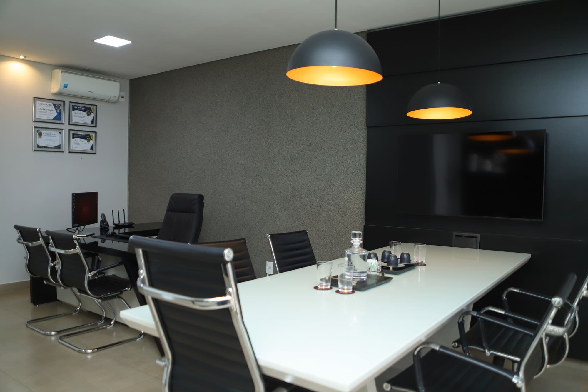 Sala de reuniões com mesa branca, cadeiras pretas e luminárias sobre a mesa